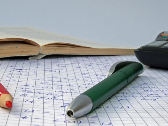 Stifte, aufgeschlagenes Buch und Taschenrechner auf beschriebenem kariertem Blatt Papier